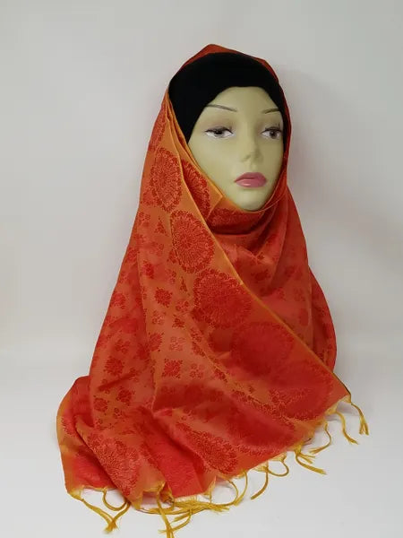 Orange and gold patterned hijab scarf with fringe edges draped on mannequin, lightweight modest wrap, soft textured fabric, full coverage stylish headscarf for everyday wear